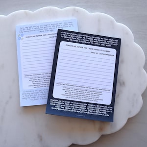May include: Two lined notepads with religious text. One notepad is light blue with floral accents, the other is black. Both have the words "Forgive me Father, for I have sinned" printed on them. The notepads are on a white, scalloped tray.