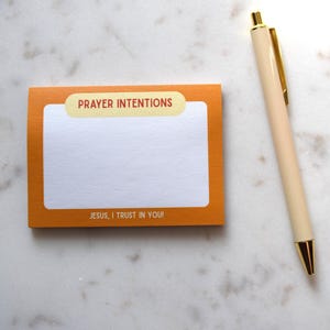 May include: An orange notepad with the words "PRAYER INTENTIONS" and "JESUS, I TRUST IN YOU!" printed on it, next to a cream-colored pen with gold accents. The notepad has a white interior for writing. The items are on a white, scalloped surface.