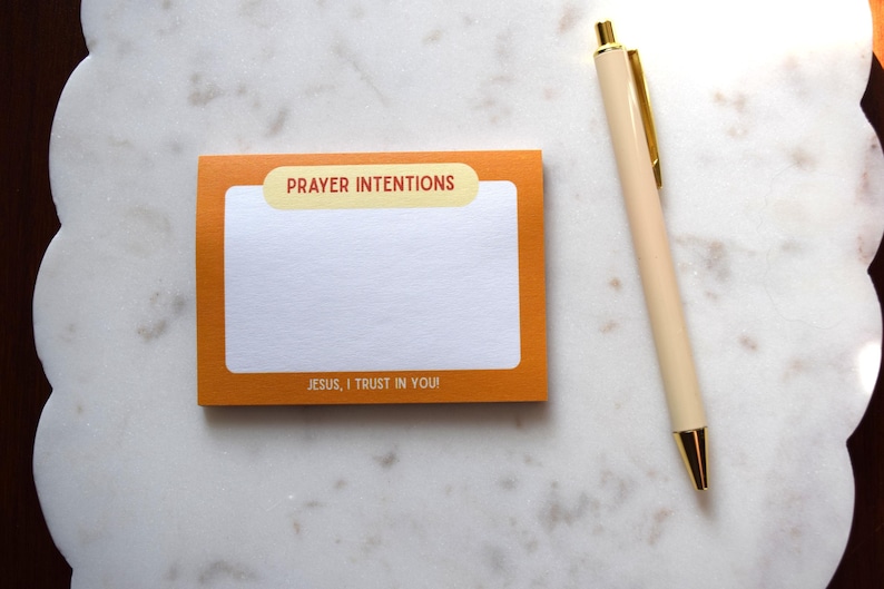 May include: An orange notepad with the words "PRAYER INTENTIONS" and "JESUS, I TRUST IN YOU!" printed on it, next to a cream-colored pen with gold accents. The notepad has a white interior for writing. The items are on a white, scalloped surface.