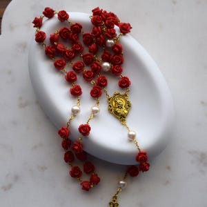 May include: A rosary with red rose beads and pearl accents. The gold-toned metal chain features a crucifix and a medallion with a religious figure. The rosary is displayed on a white, scalloped-edge dish.