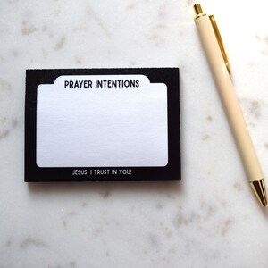 May include: A black and white prayer intentions card with the text "PRAYER INTENTIONS" and "JESUS, I TRUST IN YOU!" is next to a cream-colored pen with gold accents. The card is on a white, scalloped surface.