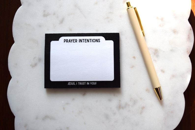 May include: A black and white prayer intentions card with the text "PRAYER INTENTIONS" and "JESUS, I TRUST IN YOU!" is next to a cream-colored pen with gold accents. The card is on a white, scalloped surface.