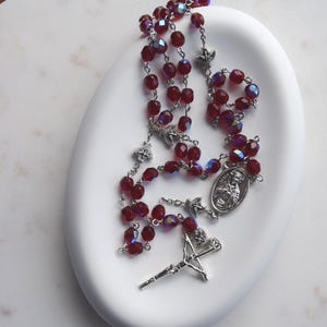 May include: A rosary with deep red, faceted beads and silver-toned metal components. The rosary features a crucifix, a center medal, and connecting chain links. The beads are arranged on a white, oval-shaped dish.