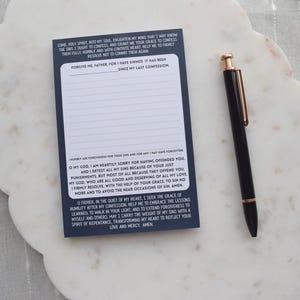 May include: A navy blue notepad with lined pages and printed text, alongside a black pen with gold accents. The notepad rests on a white, scalloped-edge tray. The text on the notepad is a prayer.