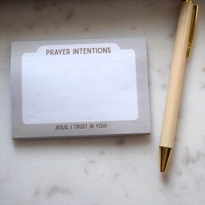 May include: A notepad with the words "PRAYER INTENTIONS" and "JESUS, I TRUST IN YOU!" printed on it, next to a cream-colored pen with gold accents. The items are on a white marble surface.