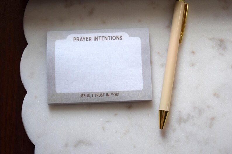 May include: A notepad with the words "PRAYER INTENTIONS" and "JESUS, I TRUST IN YOU!" printed on it, next to a cream-colored pen with gold accents. The items are on a white marble surface.
