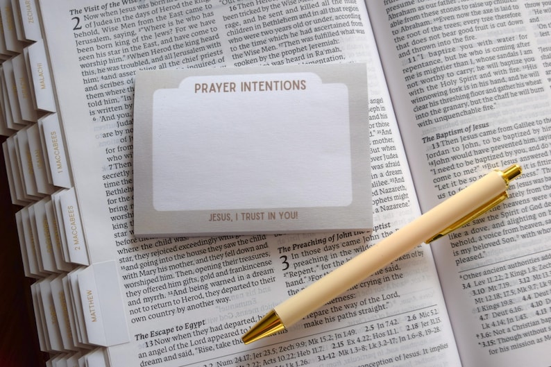 May include: A rectangular notepad with a light gray border and white center, featuring the text PRAYER INTENTIONS at the top and JESUS, I TRUST IN YOU! at the bottom, resting on an open book with a cream-colored ballpoint pen.