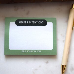 May include: A green notepad with the words "PRAYER INTENTIONS" and "JESUS, I TRUST IN YOU!" printed on it. A beige pen with gold accents rests beside the notepad on a white marble surface.