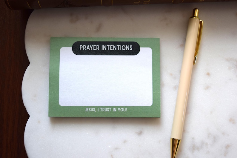 May include: A green notepad with the words "PRAYER INTENTIONS" and "JESUS, I TRUST IN YOU!" printed on it. A beige pen with gold accents rests beside the notepad on a white marble surface.