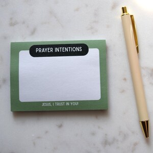 May include: A green notepad with the words "PRAYER INTENTIONS" and "JESUS, I TRUST IN YOU!" printed on it, next to a cream-colored pen with gold accents. The notepad is on a white marble surface.