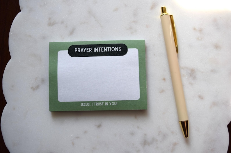 May include: A green notepad with the words "PRAYER INTENTIONS" and "JESUS, I TRUST IN YOU!" printed on it, next to a cream-colored pen with gold accents. The notepad is on a white marble surface.