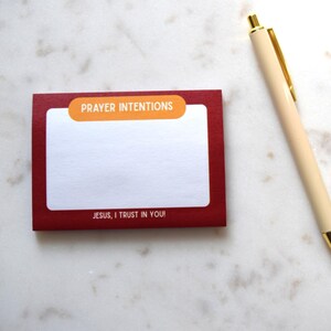 May include: A red notepad with the words "PRAYER INTENTIONS" and "JESUS, I TRUST IN YOU!" printed on it, along with a cream-colored pen with gold accents. The notepad is on a white, patterned surface.