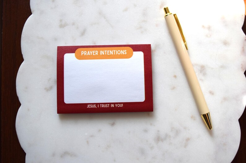 May include: A red notepad with the words "PRAYER INTENTIONS" and "JESUS, I TRUST IN YOU!" printed on it, along with a cream-colored pen with gold accents. The notepad is on a white, patterned surface.