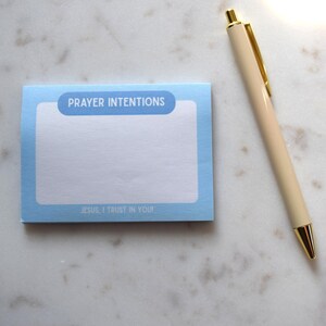 May include: A light blue notepad with the words "PRAYER INTENTIONS" and "JESUS, I TRUST IN YOU" printed on it. A beige pen with gold accents rests beside the notepad on a white marble surface.