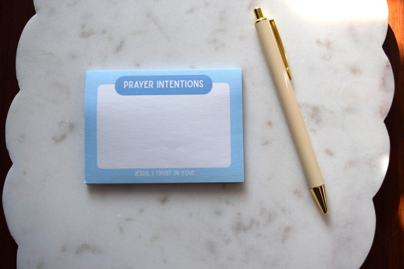 May include: A light blue notepad with the words "PRAYER INTENTIONS" and "JESUS, I TRUST IN YOU" printed on it. A beige pen with gold accents rests beside the notepad on a white marble surface.