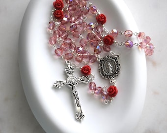 May include: A rosary with pink crystal beads and red rose accents. The rosary features a silver crucifix and a silver medallion with a religious figure. The rosary is displayed on a white, oval-shaped dish.