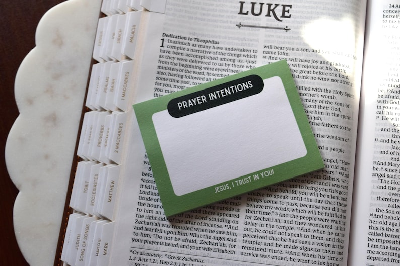 May include: Open Bible with a green and white card that reads "Prayer Intentions" and "Jesus, I trust in You!" The Bible has white tabs and is open to the book of Luke.