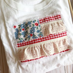 May include: A white t-shirt featuring a rectangular patch on the front. The patch is designed with a blue floral fabric section and alternating horizontal stripes of red and white gingham fabric layered with cream-colored eyelet lace ruffles.