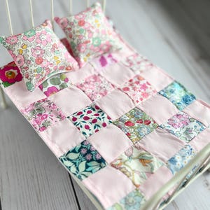 May include: Miniature white metal bed with a pink patchwork quilt and two floral print pillows. The quilt features squares of various floral fabrics and solid pink fabric. The bed frame has gold accents.