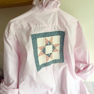 May include: A light pink button-down shirt featuring a ruffled collar and a square quilt patch sewn onto the back. The patch displays a star pattern in shades of pink, white, and dark teal with a small floral print border in dark teal.