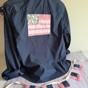 May include: A dark navy blue long-sleeve button-down shirt with a white polka dot pattern and a ruffled collar. A rectangular patch featuring a floral print and horizontal rows of white eyelet lace is sewn onto the back of the shirt.