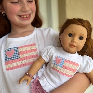 May include: A young girl holds a doll wearing a matching outfit. Both the girl and the doll are wearing white t-shirts with a pink, blue, and white flag design. The doll also wears a pink skirt and bracelet. The girl has red hair and freckles.