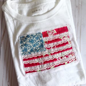 May include: A white Old Navy t-shirt featuring a rectangular applique on the front. The applique is designed like a flag with a blue floral canton and alternating stripes of pink floral fabric and white eyelet lace trim stitched onto the shirt.