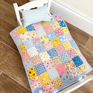 May include: A miniature white rocking chair with a patchwork quilt and pillow set. The quilt features a variety of colorful patterned squares in pink, yellow, blue, and other pastel colors. The pillow is light blue with a white floral pattern.