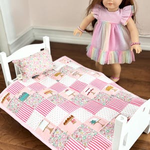 Pode incluir: Uma cama de boneca branca com uma colcha e uma almofada de patchwork rosa e branco. A colcha apresenta quadrados com motivos florais, listrados e com tema de bolo. Uma boneca com um vestido arco-íris está por perto.