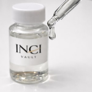 Op de afbeelding: Een heldere glazen fles met een witte dop en een label met de tekst "INCI VAULT". Een glazen pipet geeft een heldere vloeistof af, met een enkele druppel. De afbeelding is tegen een witte achtergrond geplaatst, wat het product benadrukt.
