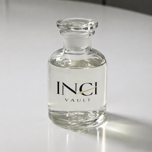Op de afbeelding: Een heldere glazen fles met een afgeronde body en een glazen stopper. De fles is gevuld met een heldere vloeistof en heeft de woorden "INCI VAULT" in zwart gedrukt met een gouden lijnaccent.