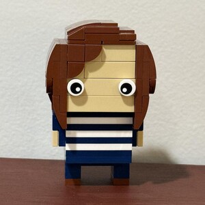 May include: A Lego figurine of a person with brown hair, a blue and white striped shirt, and blue pants.
