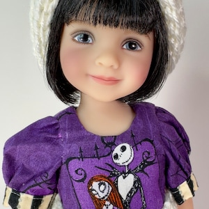 Puede incluir: Muñeca con cabello negro y ojos azules, vestida con un vestido morado con un estampado de Jack Skellington y Sally. La muñeca también lleva un gorro y un cinturón de ganchillo color crema.