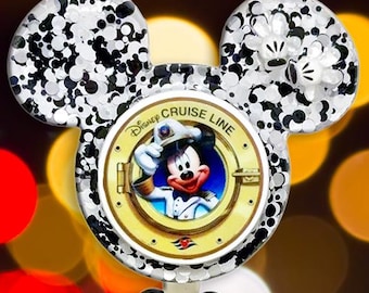 May include: A badge reel featuring a Mickey Mouse-shaped topper filled with black and white glitter. The center displays a gold porthole frame with Mickey Mouse in a captain's uniform and the text Disney Cruise Line. A black Mickey-shaped bead and patterned beads hang below.
