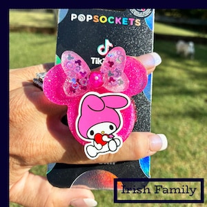 May include: A pink PopSockets phone grip featuring a Minnie Mouse-inspired design with a bow and a My Melody character holding a heart. The grip is glittery and has the TikTok logo on the phone.