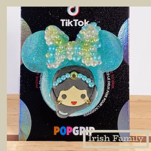 May include: A PopGrip phone grip featuring a cartoon princess design with a blue bow and a princess face. The grip is on a black background with the TikTok logo and the words "Irish Family".