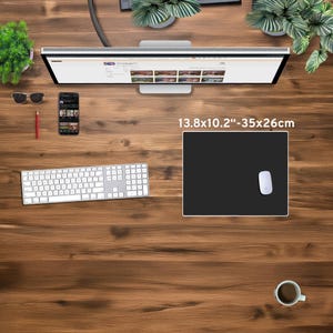 May include: A black rectangular mouse pad, measuring 13.8 x 10.2 inches (35 x 26 cm), with a white mouse on the right side. The pad is on a wooden desk, alongside a keyboard, monitor, phone, and coffee cup.