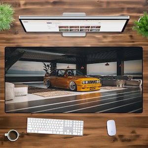 以下が含まれることがあります： 海が見えるガレージに駐車された黄色のBMW E30 M3。車は黒いホイールと黒い内装です。ガレージはコンクリートの床と白い壁です。