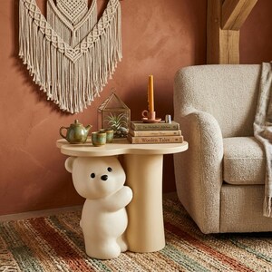 May include: A cream-colored side table with a bear-shaped base. The table has a round top with a teapot, cups, books, and a candle. The background includes a macrame wall hanging and a beige armchair.