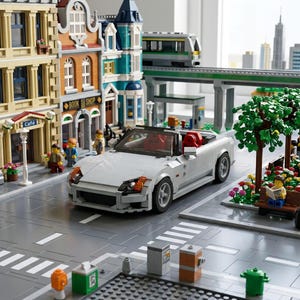 May include: A detailed Lego city scene featuring a white convertible car with red seats driving on a gray street. Buildings, a train, trees, and miniature figures add to the urban landscape. The scene includes a cafe and a bookstore.