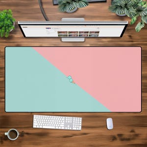May include: A large rectangular desk mat with a diagonal split design. One half is pastel pink, the other is pastel blue-green. A small illustration of two ice cream scoops on a cone is in the corner. A keyboard, mouse, and coffee cup are visible.