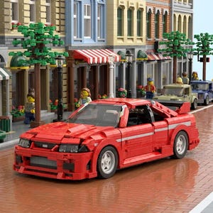 May include: A red sports car model made of interlocking toy bricks. The car has a spoiler, white racing stripes, and silver wheels. The background shows a street scene with buildings, trees, and miniature figures.