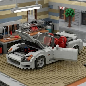 May include: A white Lego sports car with the hood and door open, revealing a red interior and engine components. The car is in a Lego garage setting with tools and storage. The scene includes a workbench, tools, and a street scene in the background.