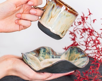 May include: A ceramic mug and matching dish set. The mug has a brown rim and handle, with a cream and brown glaze. The dish has a wavy shape with a similar glaze and a dark rim. The set is handmade and suitable for serving beverages and snacks.
