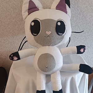 May include: A white and gray plush cat toy with black ears, paws, and tail. The cat has large black eyes and a pink nose.