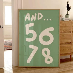 May include: A framed art print with a light green background and the words "AND..." above the numbers "5678" in white. The print is displayed on a wooden floor, leaning against a wall. A wooden cabinet and a potted plant are also visible.