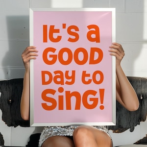 Op de afbeelding: Een roze ingelijste poster met de tekst "It's a good day to sing!" in oranje letters. De poster wordt vastgehouden door een persoon die op een zwart-witte koe-printbank zit.