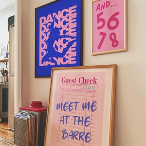 May include: Three framed prints on a white wall. The leftmost print is blue with a pink silhouette of a dancer and the text "DANCE" repeated multiple times. The middle print is pink with the text "AND... 56 78". The rightmost print is pink with a white check-like design and the text "Guest Check" and "MEET ME AT THE BARRE".