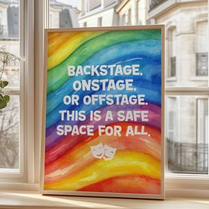 May include: A framed print with a rainbow watercolour background and white text that reads "BACKSTAGE, ONSTAGE, OR OFFSTAGE. THIS IS A SAFE SPACE FOR ALL." Two white theatre masks are at the bottom of the print.