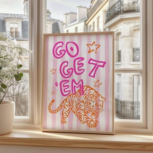 K&ouml;nnte beinhalten: Ein gerahmter Kunstdruck mit dem Text "GO GET 'EM" in Pink, mit orangefarbenen Sternen und einer Tigerillustration. Der Hintergrund hat rosa und wei&szlig;e vertikale Streifen. Das Kunstwerk wird auf einer Holzfensterbank ausgestellt.